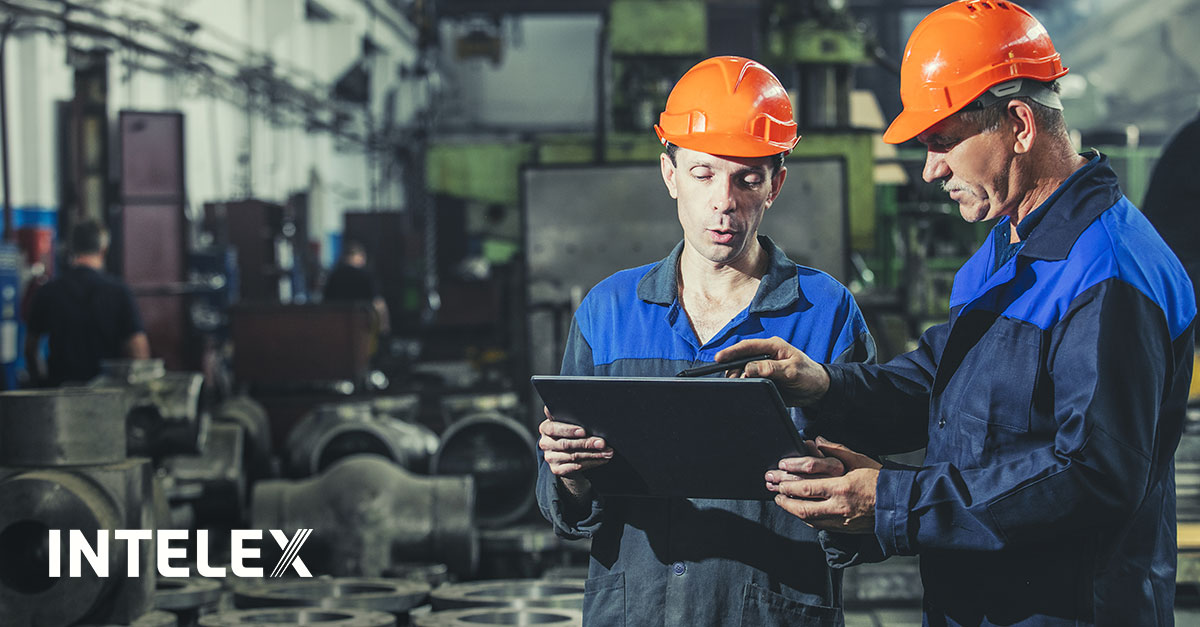 Two factory workers in safety gear review data on a tablet